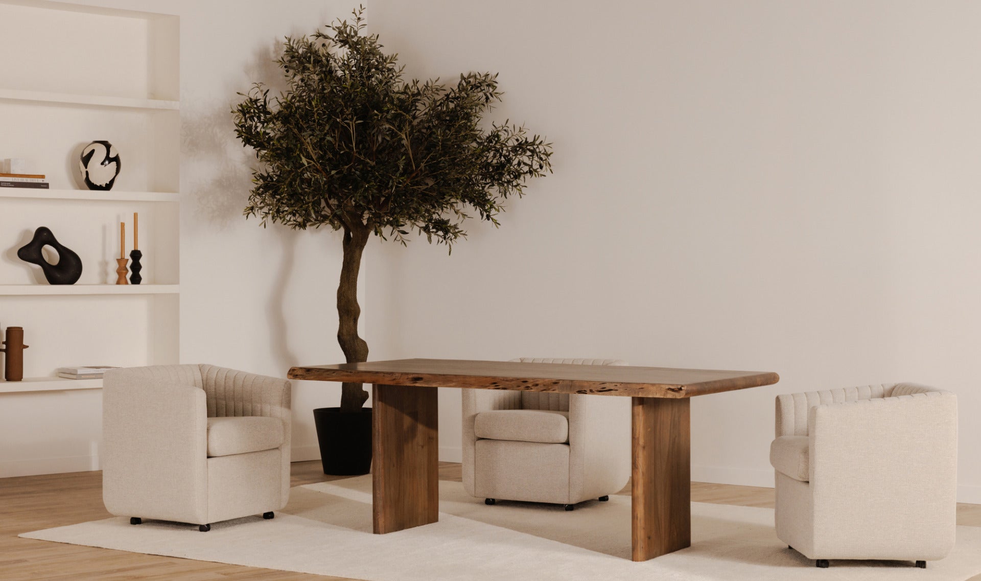 A modern living room scene featuring a rectangular wooden dining table with thick block legs, surrounded by three cream-colored armchairs and a large potted olive tree against a warm beige wall.