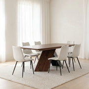 A modern dining table set in a bright, minimalist room. The set includes a rectangular dark wood dining table with a sculptural angled pedestal base, surrounded by six white upholstered dining chairs with slim black metal legs, all arranged on a textured cream-colored area rug.