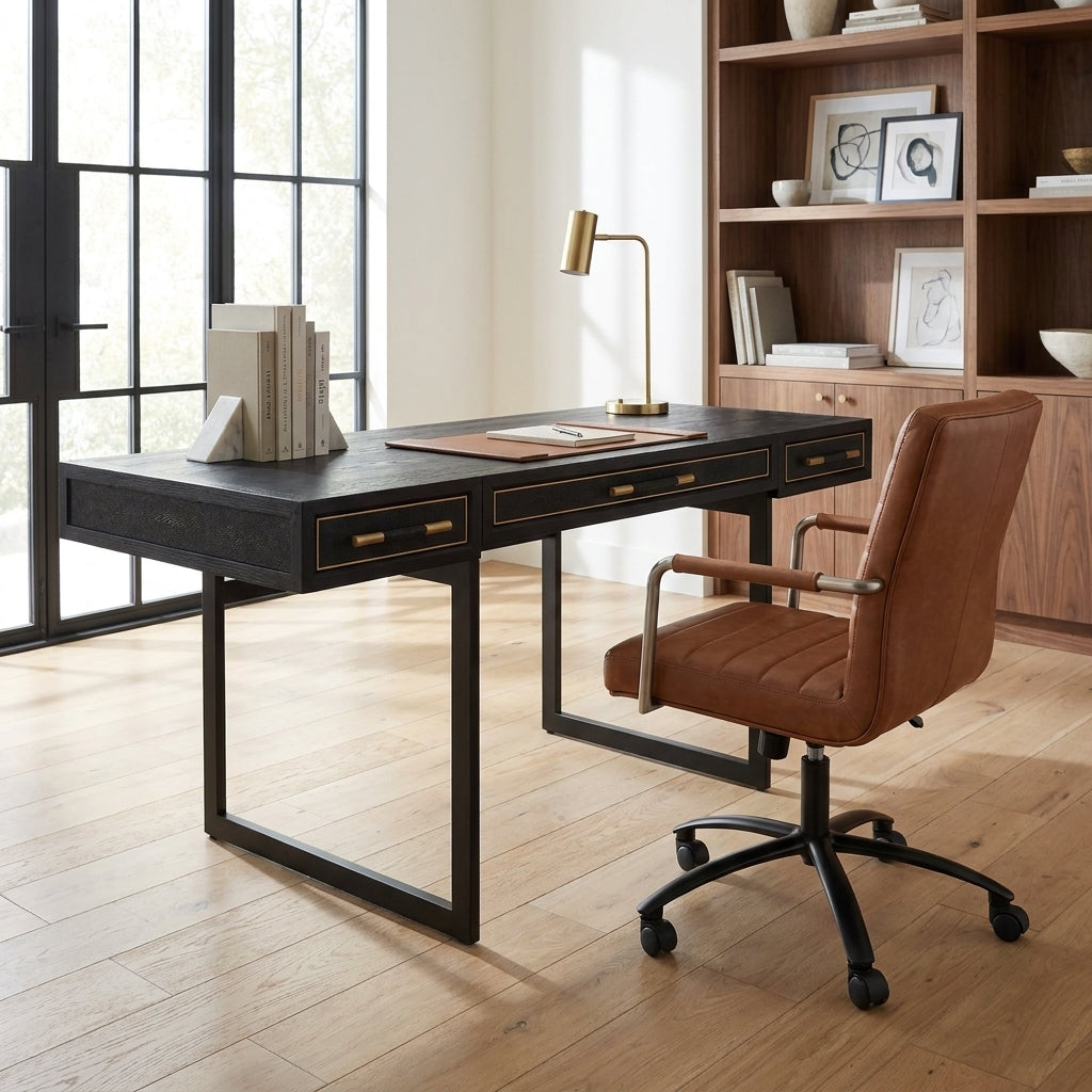A modern home office setup featuring a black wood writing desk with brass hardware, a matching black metal desk chair with a brown leather seat, and a brass desk lamp, set in a bright room with light wood floors and a wooden bookshelf.