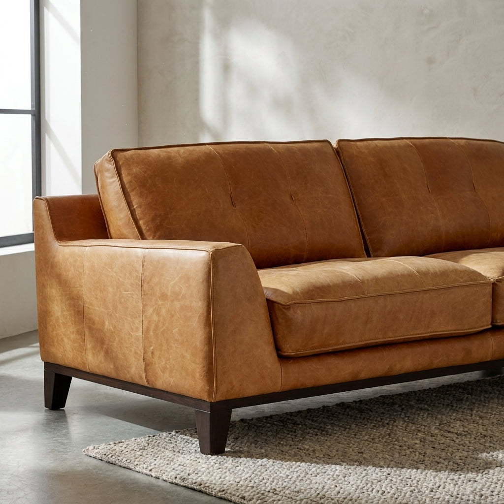 A modern two-seater sofa upholstered in warm tan leather with dark wooden legs, shown in a minimalist room setting with a textured rug and concrete floor.