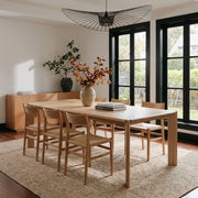 A light natural wood dining table with six matching wooden chairs in a bright, modern dining room with black-framed windows and a woven rug.