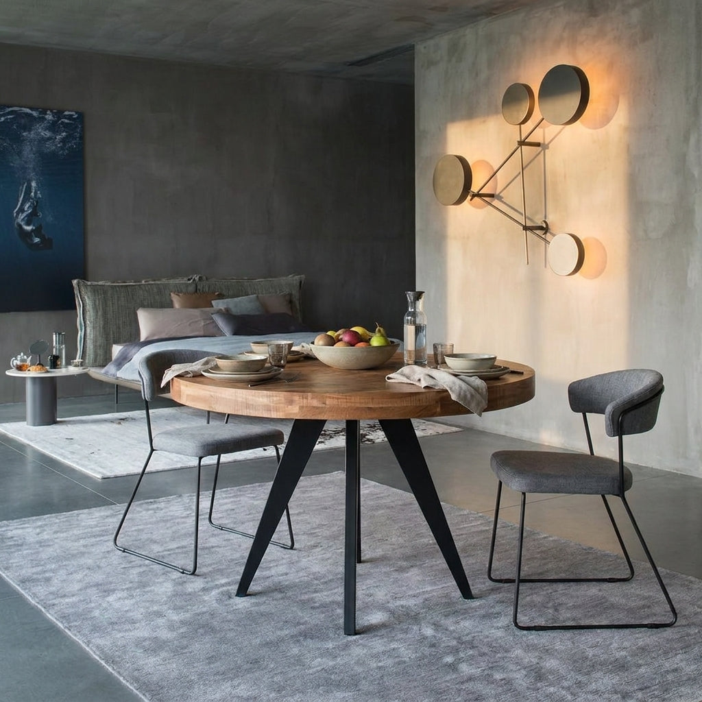 A round wooden dining table with black metal legs is styled in a modern dining room setting with gray upholstered chairs, a gray rug, and a concrete wall with a geometric wall light.