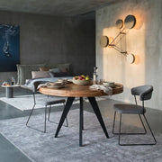 A round wooden dining table with black metal legs is styled in a modern dining room setting with gray upholstered chairs, a gray rug, and a concrete wall with a geometric wall light.