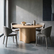 A round light oak dining table with a sculptural wavy pedestal base, surrounded by four gray upholstered dining chairs in a modern minimalist dining room with concrete floors and walls.