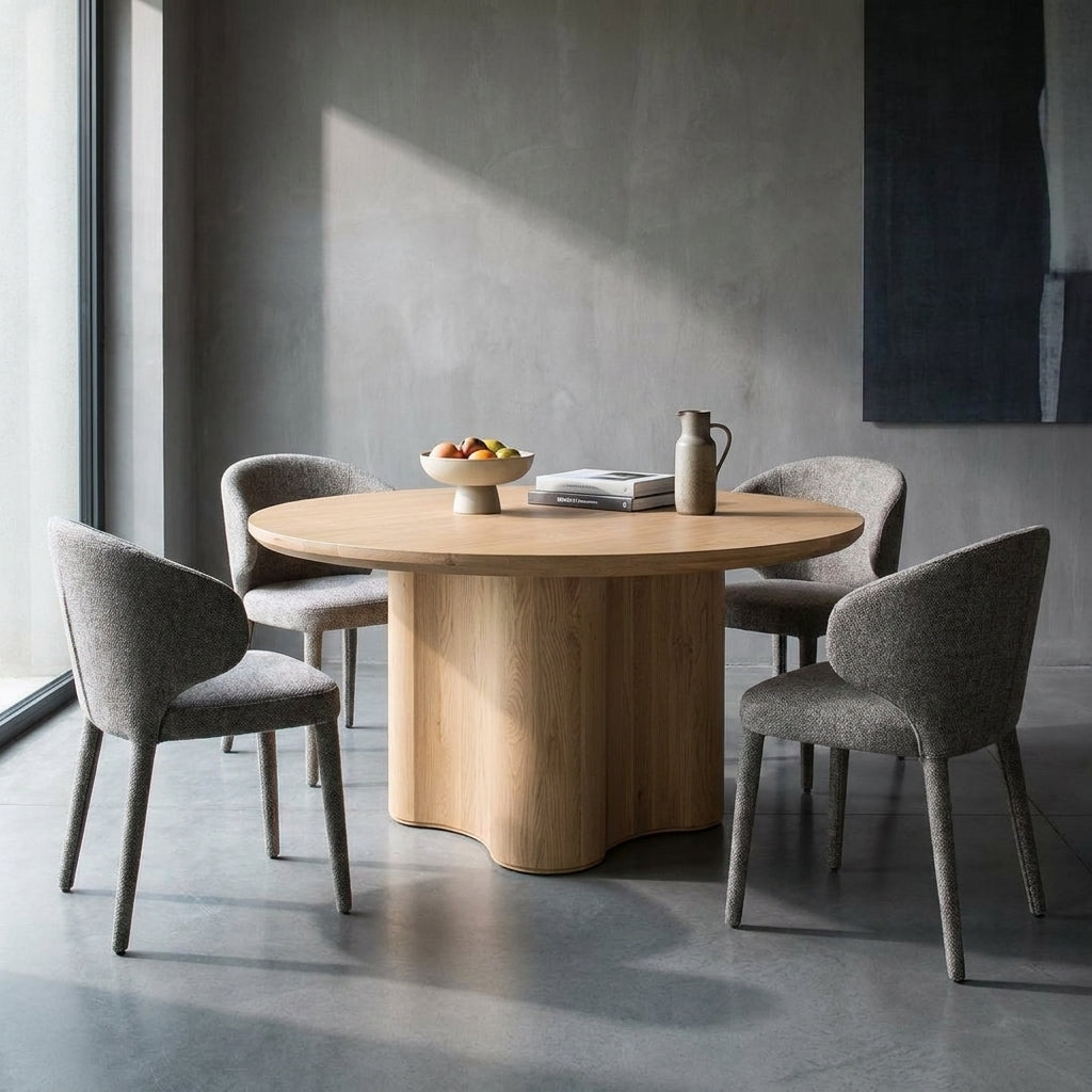 A round light oak dining table with a sculptural wavy pedestal base, surrounded by four gray upholstered dining chairs in a modern minimalist dining room with concrete floors and walls.