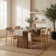 A rustic wooden dining table with four matching wooden chairs in a warm, sunlit room. The table has a thick, live-edge wood top and a solid block base, while the chairs have woven cane seats.