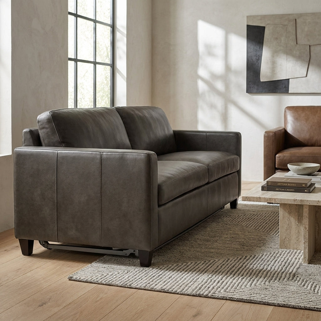 A dark brown leather loveseat sofa in a modern living room setting with a wooden floor, textured rug, and a matching brown leather armchair in the background.