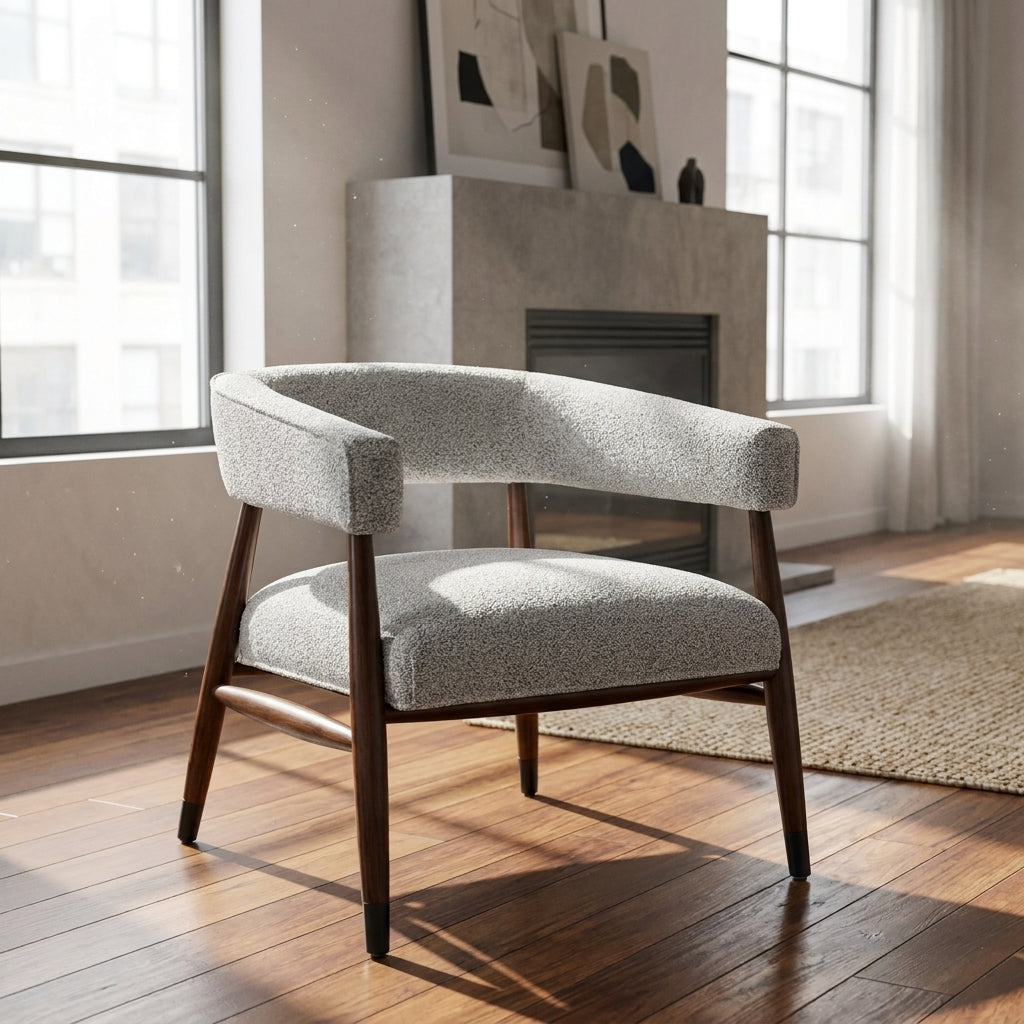 A modern accent chair with a curved backrest, upholstered in a textured light gray fabric, supported by dark brown wooden legs, set in a bright room with hardwood floors and a fireplace.