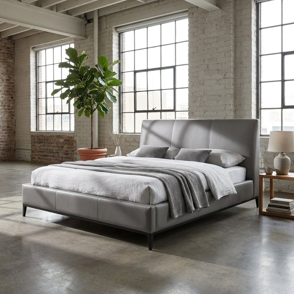 Gray upholstered bed in a room with large windows and brick walls.