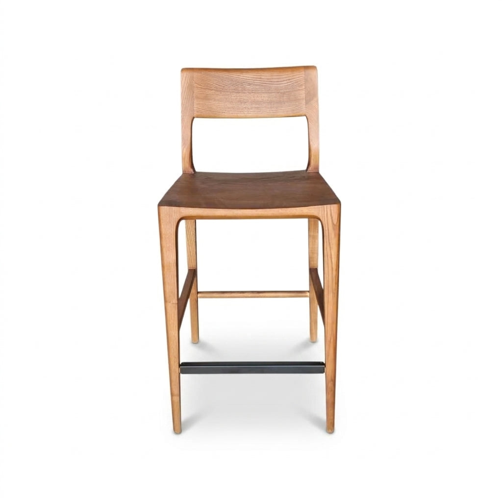 A modern wooden bar stool with a warm medium-brown finish, featuring a backrest with a central cutout, a square seat, and a black metal footrest bar, shown against a plain white background.