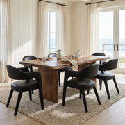 A dining set consisting of a rectangular rustic wooden table and six black upholstered chairs, styled on a woven jute rug in a bright, modern room with ocean views.