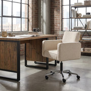 A modern upholstered office chair in a light beige textured fabric with a black swivel base and casters, positioned next to a rustic wooden desk in a loft-style office with brick columns and large windows.