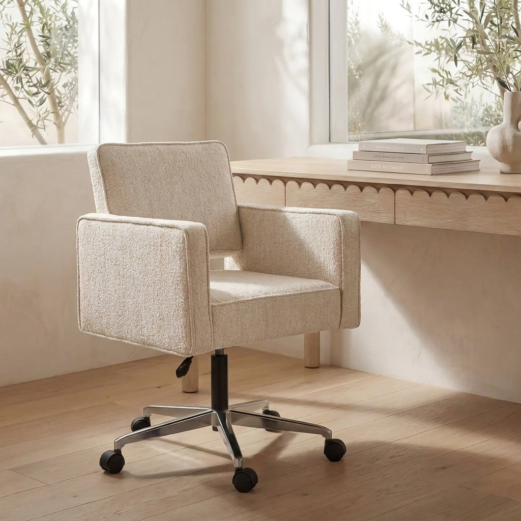 A modern upholstered office chair in a textured cream bouclé fabric, positioned in a bright, minimalist room with a wooden desk and windows in the background.