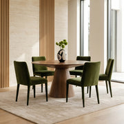 A modern dining set featuring a round wooden table and four olive green velvet chairs, arranged on a light beige rug in a contemporary dining room with wood slat walls and large windows.