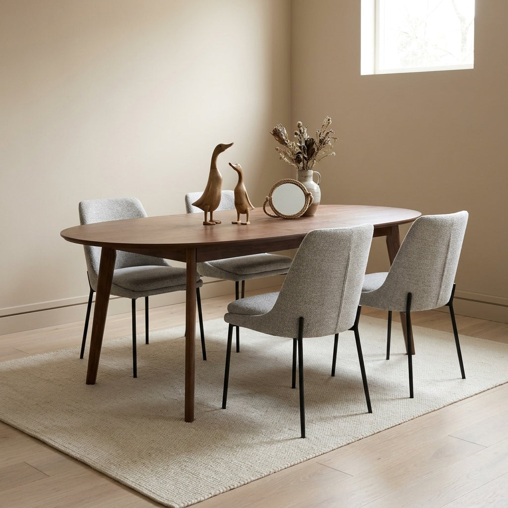 An oval wooden dining table with four upholstered gray chairs in a bright, modern dining room setting with a beige rug and light wood floor.