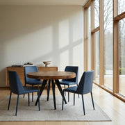 A round wooden dining table with black angled legs, surrounded by four dark blue upholstered chairs, set on a light gray rug in a bright room with large windows and natural light.