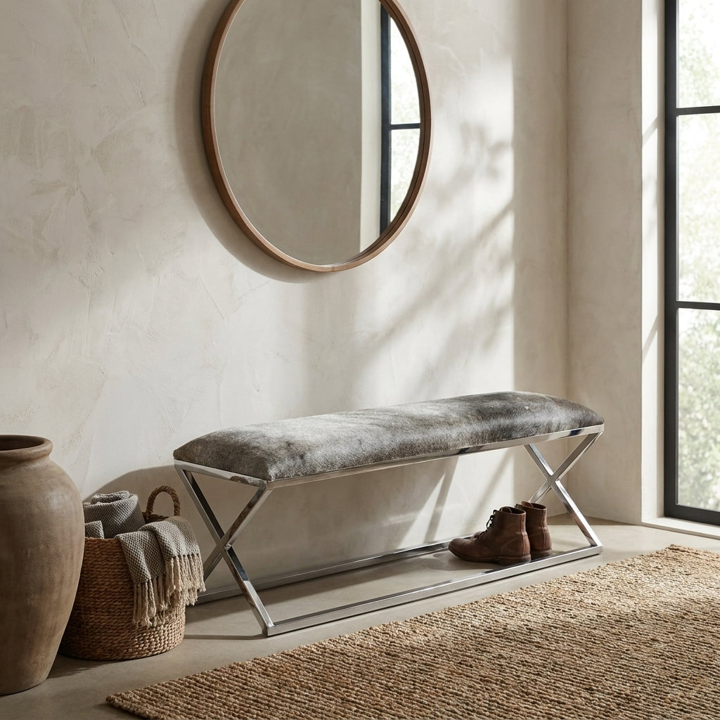 A modern bench with a gray textured faux-fur cushion and a chrome X-frame base, styled in a sunlit entryway with a round wood-framed mirror, woven baskets, and a rug.