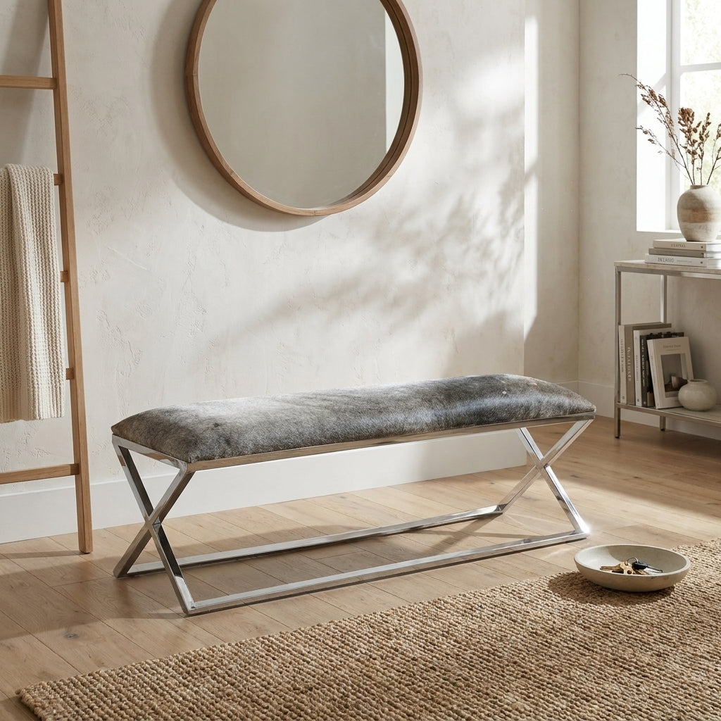 A modern entryway bench with a gray textured faux-fur cushion and a chrome X-frame base, placed in a bright, sunlit room with a round wooden mirror, wood floor, and woven rug.