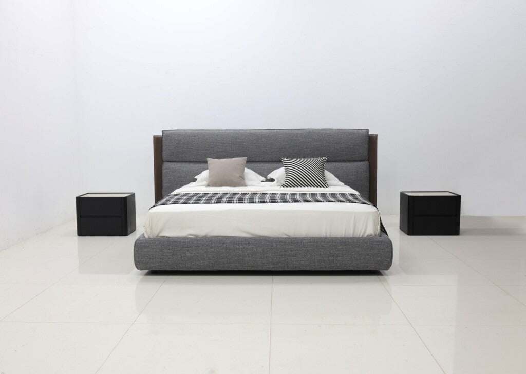A modern platform bed with a gray upholstered frame and headboard, styled with white bedding, two gray pillows, and a striped throw blanket, flanked by two black nightstands on a white tiled floor against a white wall.