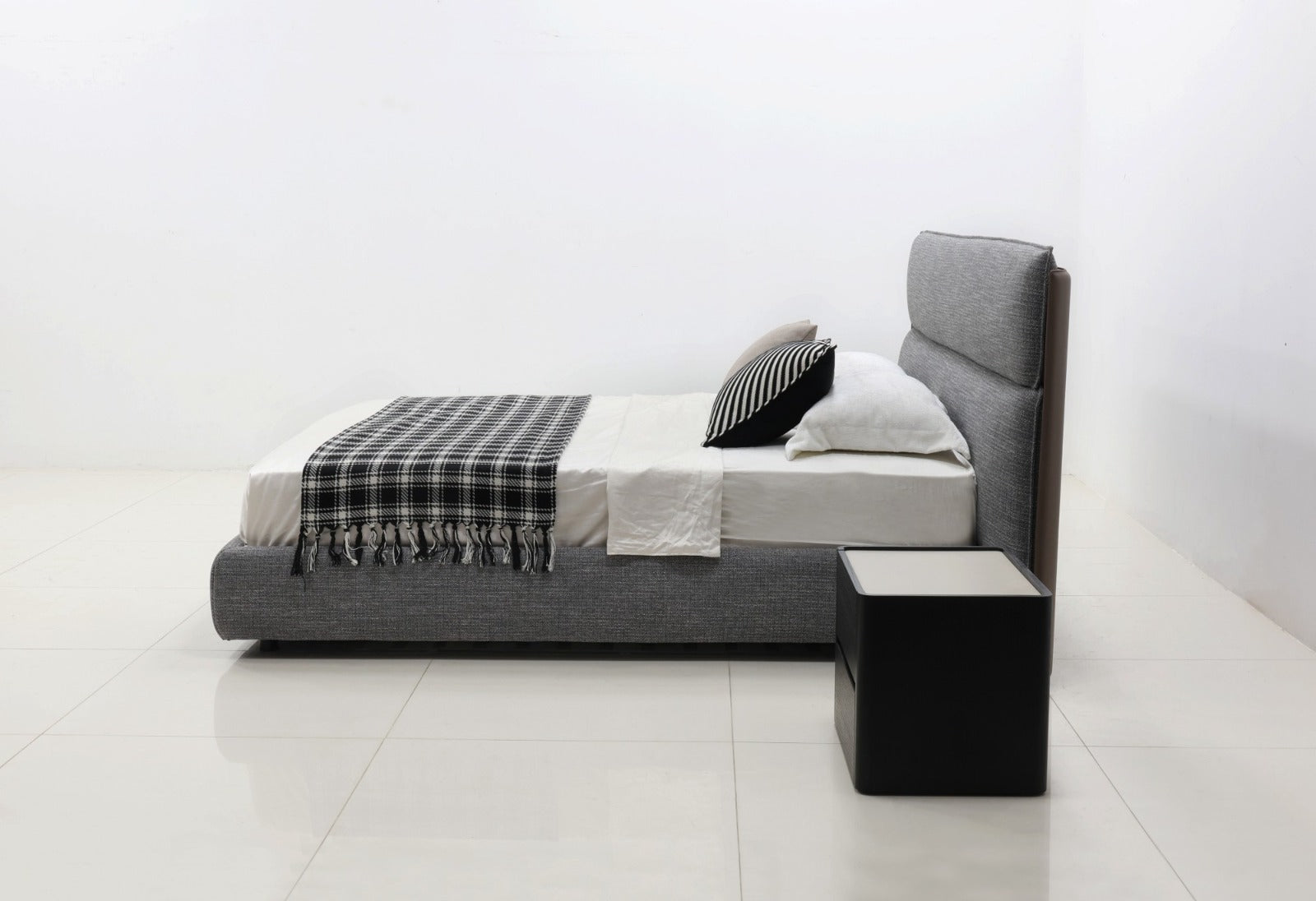 A modern low-profile platform bed with a gray upholstered headboard and base, styled with white bedding, a black-and-white plaid throw blanket, and two pillows. A matching black bedside table is positioned next to the bed in a bright, minimalist room with white walls and tiled floors.