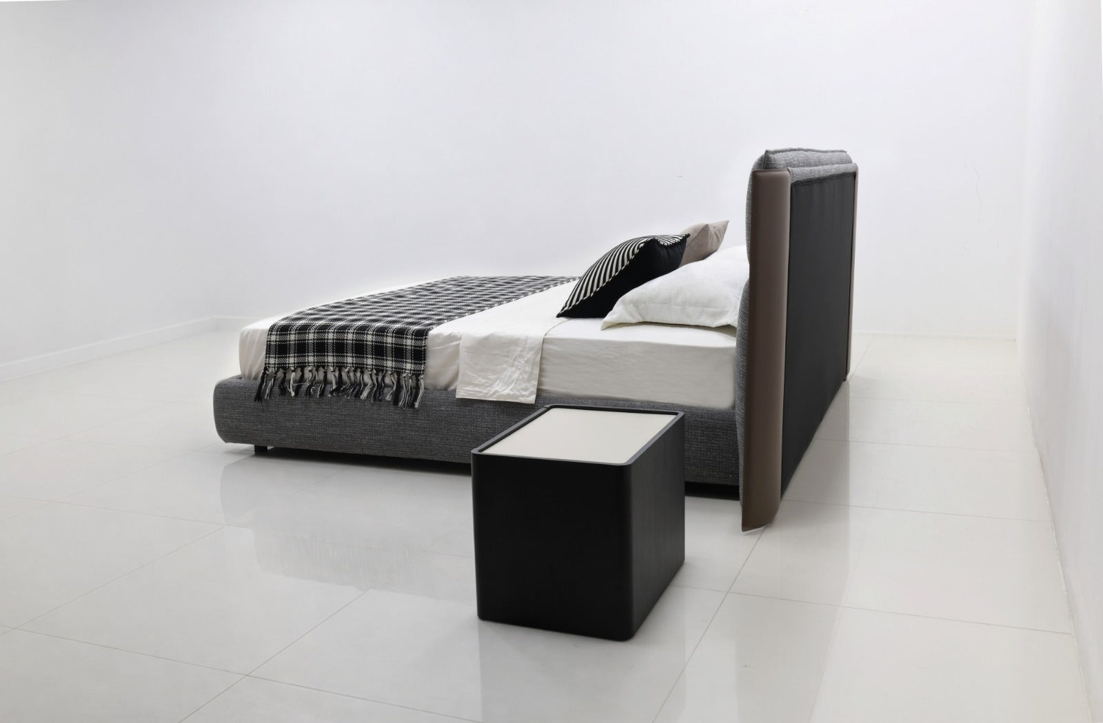 A modern platform bed with a tall upholstered headboard and matching footboard in dark gray and black, styled with white bedding and a black-and-white plaid throw blanket. A matching black cube side table sits in front of the bed in a bright, minimalist white room.