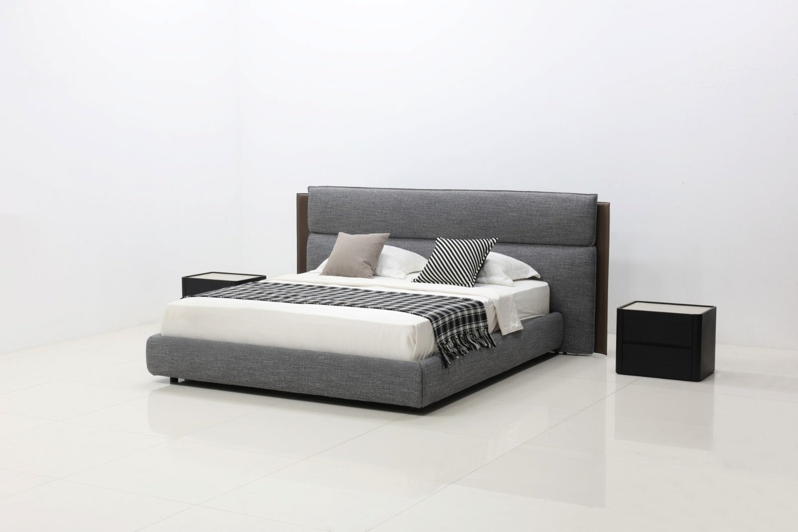 A modern platform bed with a gray upholstered frame and headboard, styled with white bedding, gray and black accent pillows, and a black-and-white striped throw blanket. Two black nightstands flank the bed on a clean white studio background.