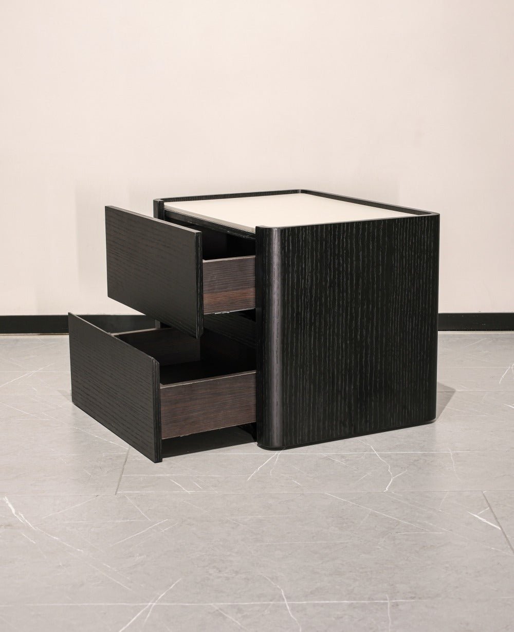 A modern black nightstand with a textured vertical wood grain finish, featuring two open drawers and a white top surface, set against a neutral light gray wall and floor.