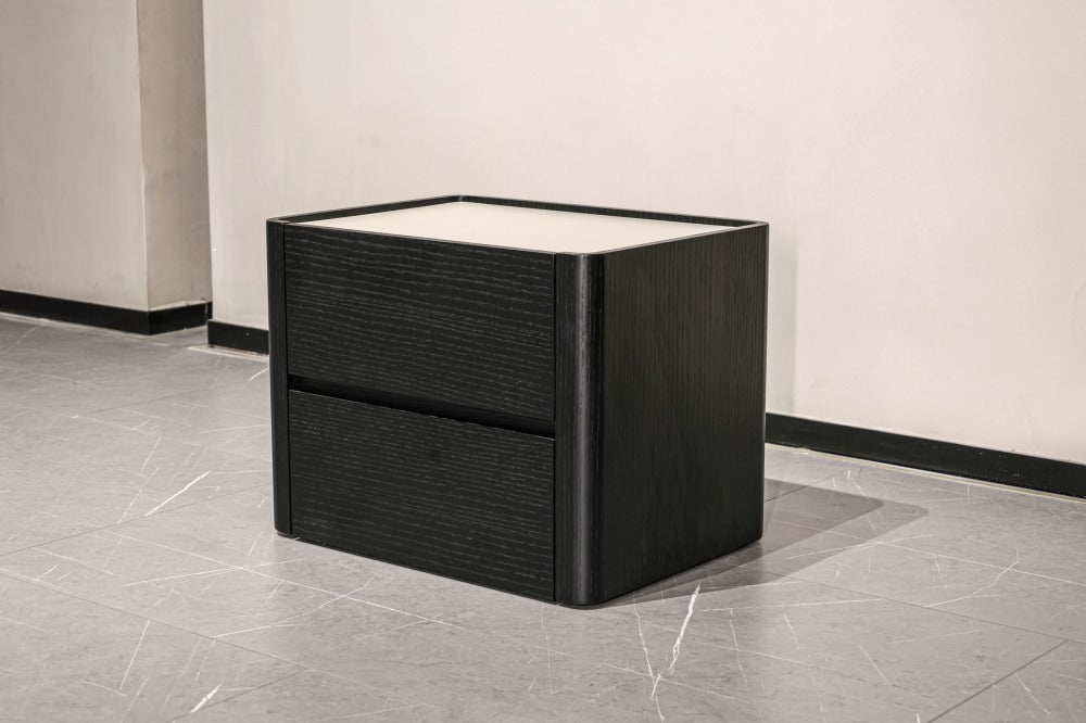A modern black two-drawer nightstand or side table with a textured wood grain finish and a light top surface, shown in a minimalist interior setting with a gray tiled floor and beige walls.